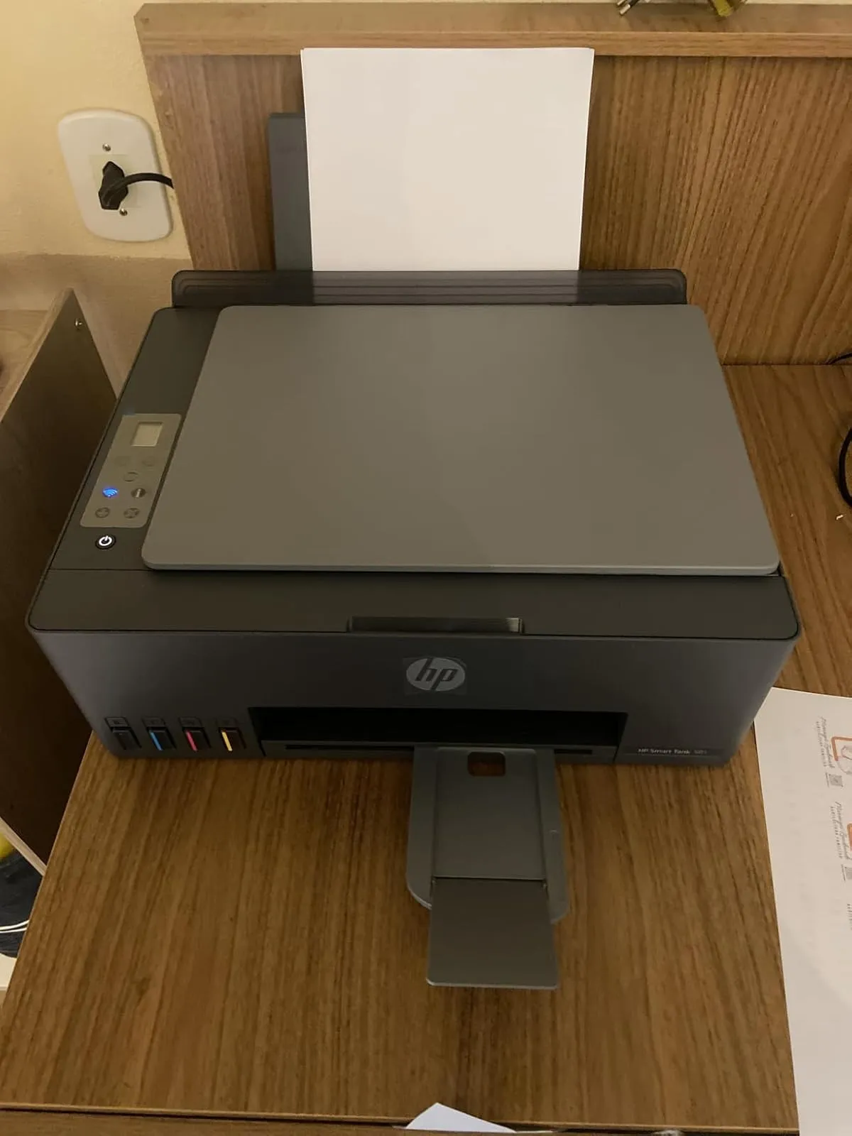 Impressora HP Smart Tank 581 ligada, com papel na bandeja de entrada e posicionada sobre uma mesa de madeira.